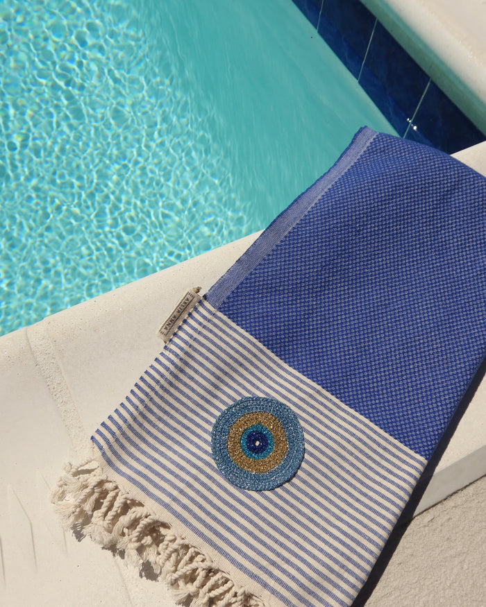 Blue and off-white striped cotton pestemal towel with an embroidered metallic evil eye and fringe trim made in Greece, Greek beach towel