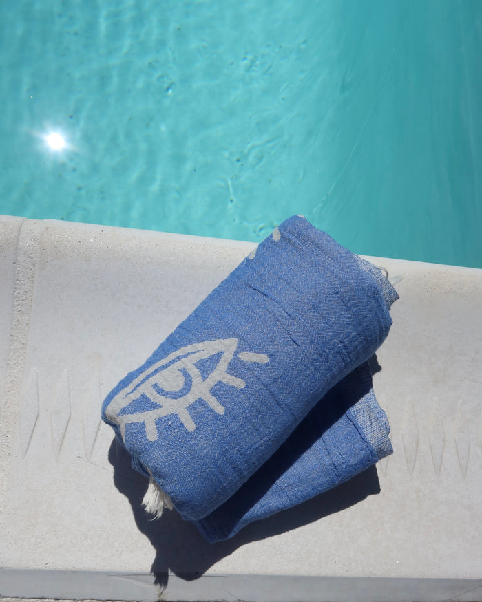 Blue cotton pestemal towel with a white evil eye print on a white surface near a pool.