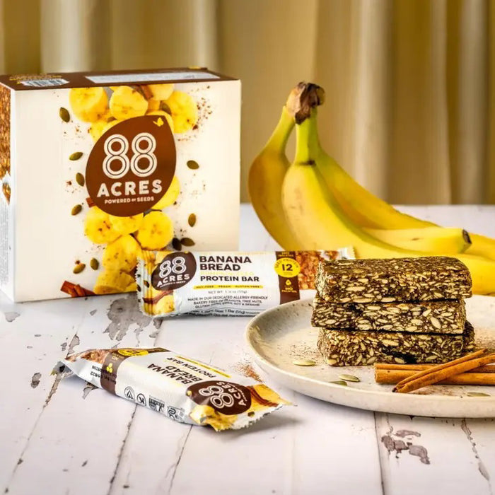 The 88 ACRES - BANANA BREAD HIGH PROTEIN BAR is shown wrapped, with packaging featuring cinnamon sticks, banana slices, and seeds. This nut-free bar is labeled "12g protein," "vegan," "gluten-free," and "no palm oil.