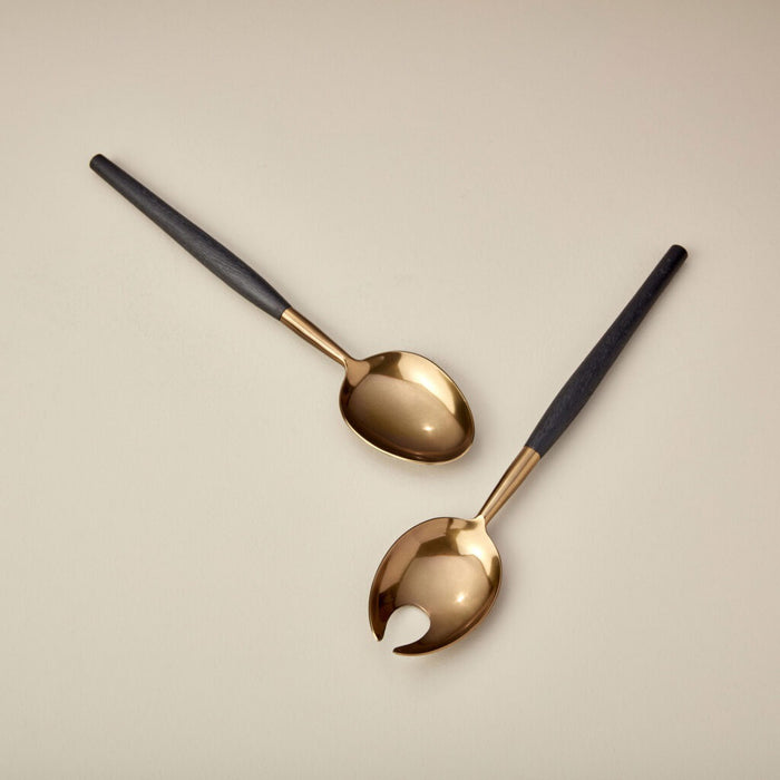 A round, deep bowl from BE HOME, INC's AGED BRONZE SERVING SET with a smooth, golden-bronze finish sits on a light wooden surface. The slightly irregular, organic shape of the bowl enhances its handcrafted appearance. The background displays the light wood grain pattern of the tabletop.