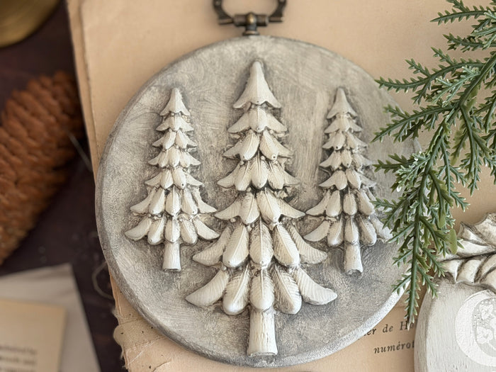 A round gray decorative plaque featuring three raised white pine tree silhouettes, styled on a beige surface with a pinecone and greenery nearby.