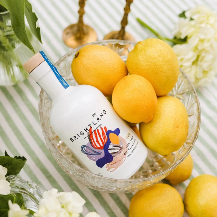 A white bottle of BRIGHTLAND ‘LUCID’ Lemon Infused Olive Oil with a wooden cap and a colorful abstract label. Crafted on California’s Central Coast for vibrant hair, this cold-pressed oil features zesty lemon flavor.