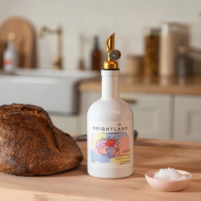 A white bottle of BRIGHTLAND 'ROSETTE' Garlic Infused Olive Oil with a wooden cap and a minimalist label featuring abstract line art, made from California olives.