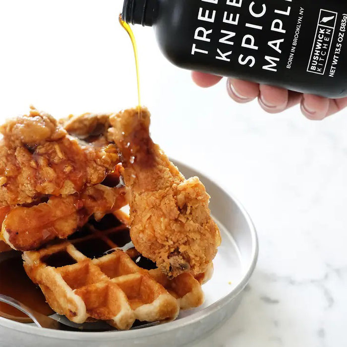 A 12.5 oz (354g) black bottle of BUSHWICK KITCHEN - 'TREES KNEES' SPICY MAPLE SYRUP features white text, a Brooklyn skyline graphic, and a black cap, containing habanero-infused spicy maple syrup.