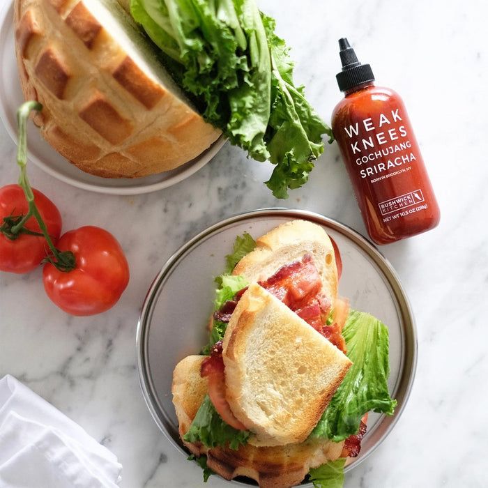 A clear bottle of BUSHWICK KITCHEN - 'WEAK KNEES' GOCHUJANG SRIRACHA hot sauce with a black squeeze cap, vibrant red chile paste, and label showing the name, "Born in Brooklyn, NY." Net wt. 10.5 oz (298g).