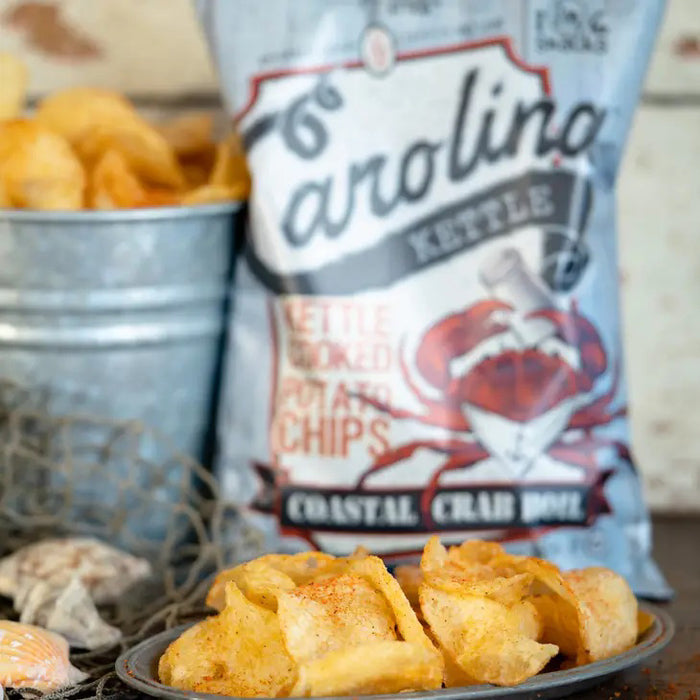 A 5oz bag of CAROLINA KETTLE - CRAB BOIL CHIPS by 1 IN 6 SNACKS features a red crab with a scroll on a blue wooden background, accented by bold red and white text.