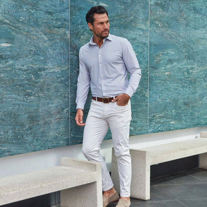 A man with short brown hair and a beard, wearing the COLLARS AND CO Semi Spread Collar Apex Performance Dress Shirt in Tan and Blue Tattersall, sits on a bench with his hands clasped, looking to the side by a blue textured wall.