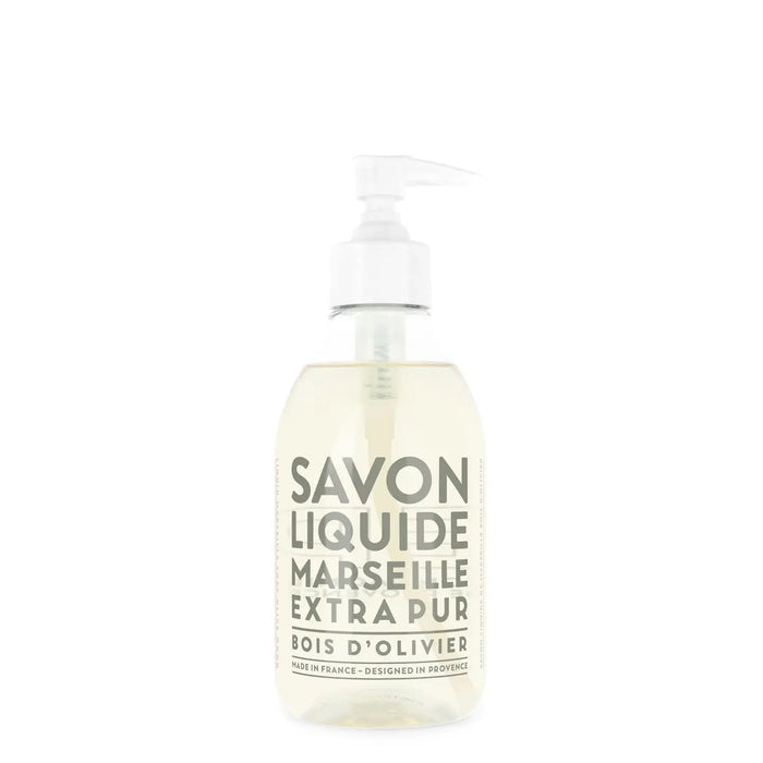 A clear pump bottle of COMPAGNIE DE PROVENCE Liquid Marseille Soap Olive Wood 10oz by CIE LUXE BRANDS, with a light-colored soap and a white pump dispenser. White label text reads "Savon Liquide Marseille Extra Pur Bois d’Olivier.