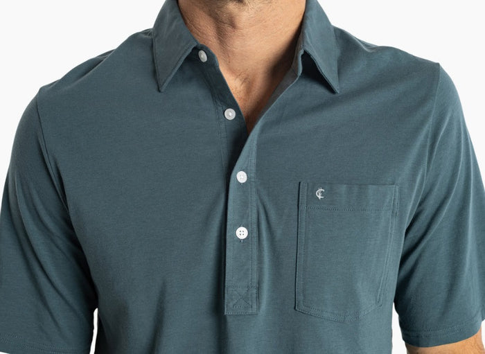 A smiling man with short dark hair wears the CRIQUET Top-Shelf Players Shirt in Dark 'n Stormy with a chest pocket and dark jeans, standing against a plain white background.
