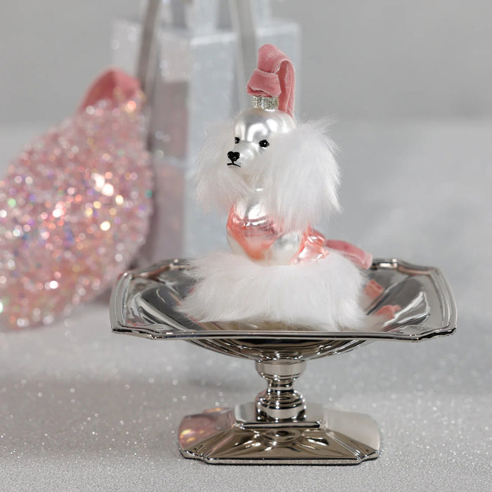 The ZODAX GLASS STANDARD POODLE ORNAMENT features a white poodle with fluffy fur and a pink outfit, hanging from a gold string. Sized 3" x 2" x 4.5", it fits perfectly in a small box—ideal for festive decor or gifting.