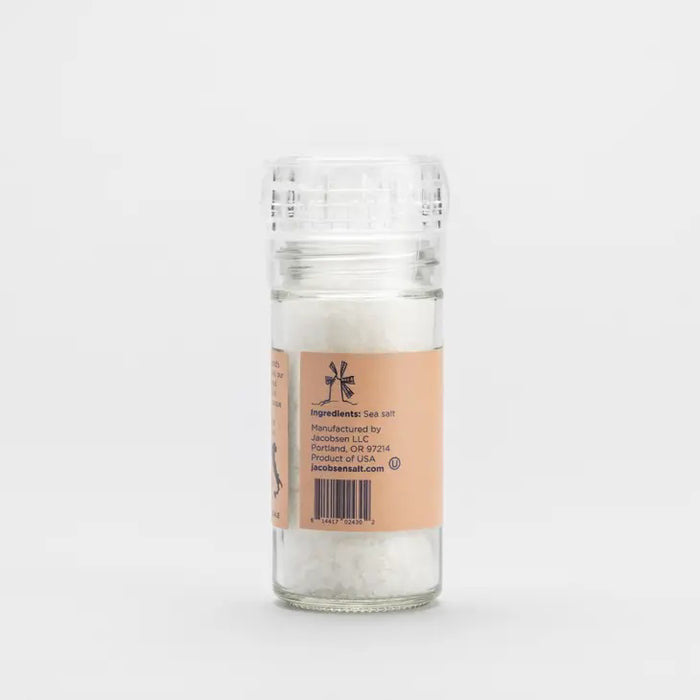 A clear plastic grinder of JACOBSEN SALT CO. JACOBSEN SALT - COARSE ITALIAN SEA SALT WITH GRINDER, sourced from the renowned Trapani region, rests on a light stone surface against a plain gray background.