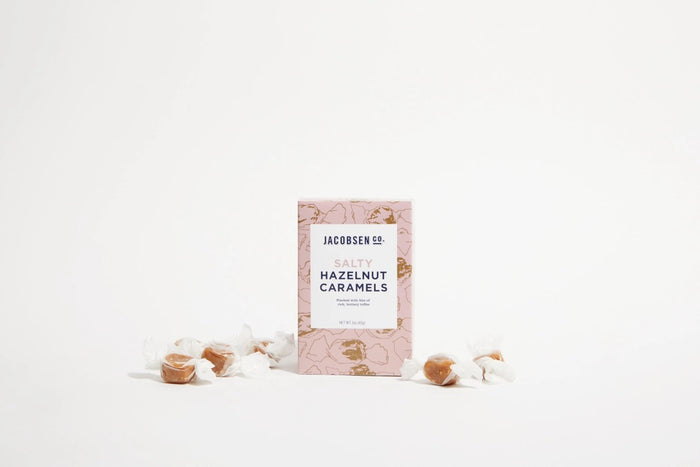 A pink JACOBSEN SALT - SALTY HAZELNUT CARAMELS BOX by JACOBSEN SALT CO. sits on a light textured surface, featuring a modern design with subtle hazelnut patterns and hints of buttery toffee made from premium hazelnuts.