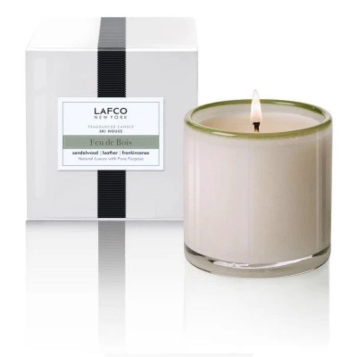 A lit LAFCO Ski House Feu de Bois candle in art glass sits next to a white LAFCO New York box. Enjoy notes of sandalwood, leather, and frankincense—a classic luxury scent.