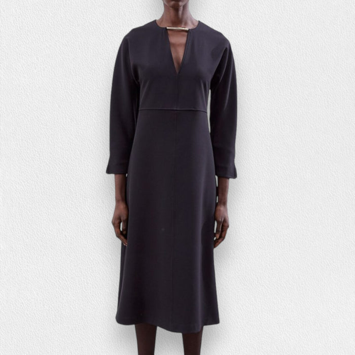 A model wears the MEIMEIJ Long Sleeve Dress with Metallic Collar—a black, midi-length dress with a V-neckline by MEIMEIJ—standing against a textured light background.