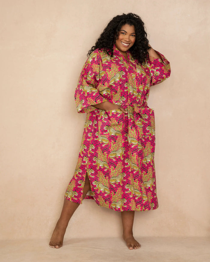 A smiling person with long hair wears the PRINTFRESH - BAGHEERA COTTON ROBE IN HOT PINK, hand screen-printed by PRINTFRESH, standing barefoot on a wooden floor against a plain light-colored wall.
