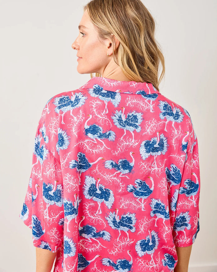 A woman with wavy blonde hair stands barefoot against a white wall, smiling softly as she models the PRINTFRESH - OSTRICH DANCE SATIN WILDEST DREAM PAJAMA SET, a matching pink pajama set by PRINTFRESH featuring a blue and white floral pattern. This eco-friendly satin sleepwear combines stylish design and sustainable comfort for your wildest dreams.