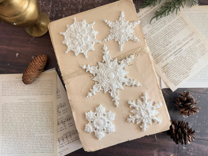 Five white lace snowflake embellishments arranged on a beige book, surrounded by vintage book pages, pinecones, and a gold goblet on a dark wooden surface.