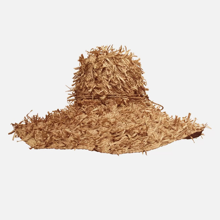 The SUMMERTIME HAT by SANS ARCIDET is a wide-brimmed, floppy raffia hat from Madagascar, featuring a rough, shaggy texture and uneven edges, shown against a plain white background.