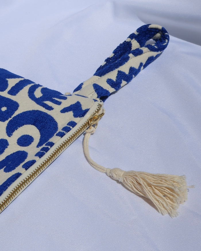 Wristlet clutch bag with a cream background and woven blue geometric symbols design with an evil eye, rainbow, moons, etc. with a gold zipper and tassel and wrist strap