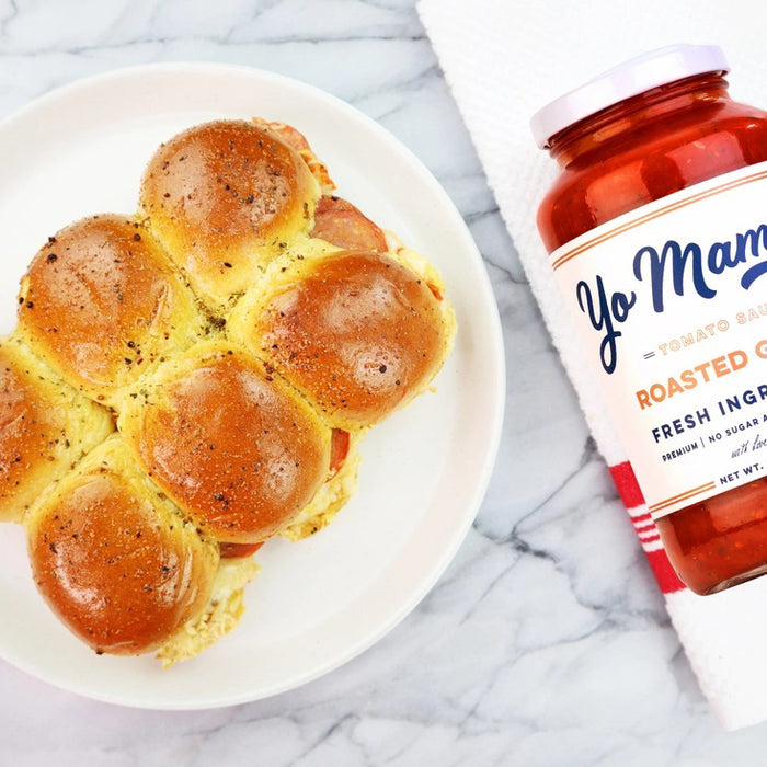 A 25 oz (708g) glass jar of YO MAMA'S FOODS ROASTED GARLIC SAUCE with a white label displaying the brand, flavor, keto certified, no sugar added, ingredients highlight; red sauce visible through the jar.