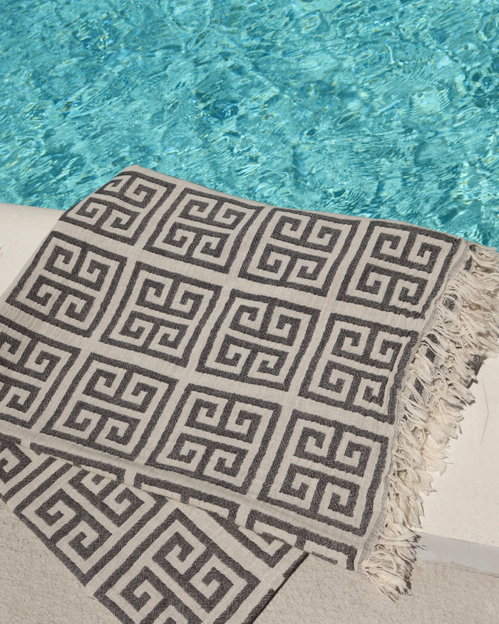 Black and off-white Greek Key meander print cotton towel next to a pool. Greek pestemal towel with fringe