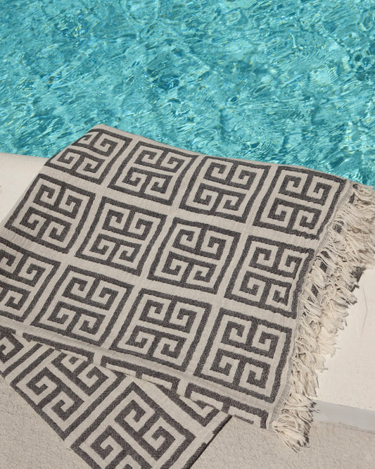 Black and off-white Greek Key meander print cotton towel next to a pool. Greek pestemal towel with fringe