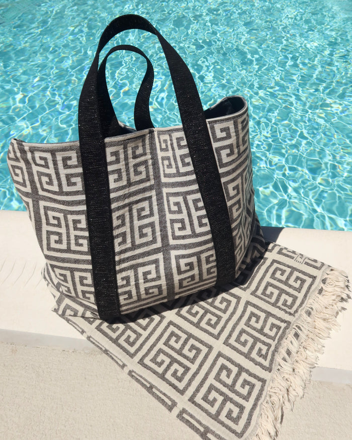 Black and off-white Greek Key meander print cotton towel and matching beach bag next to a pool. Greek pestemal towel with fringe