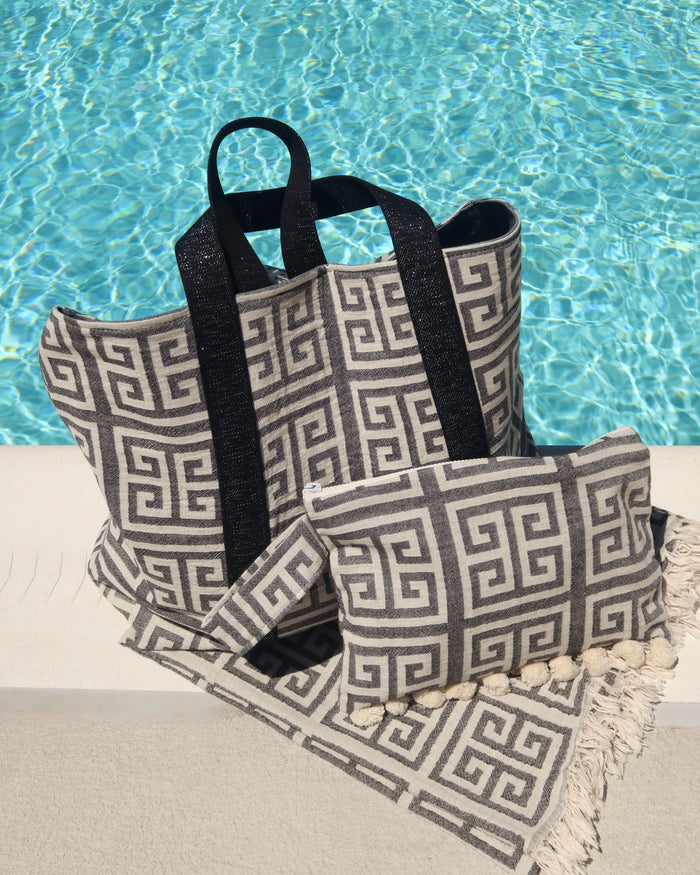 Black and off-white Greek Key meander print cotton towel, wet/dry zippered pouch clutch bag, and matching beach bag next to a pool. Greek pestemal towel with fringe