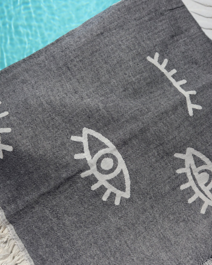 Dark gray/black cotton pestemal towel with white evil eye print next to a pool