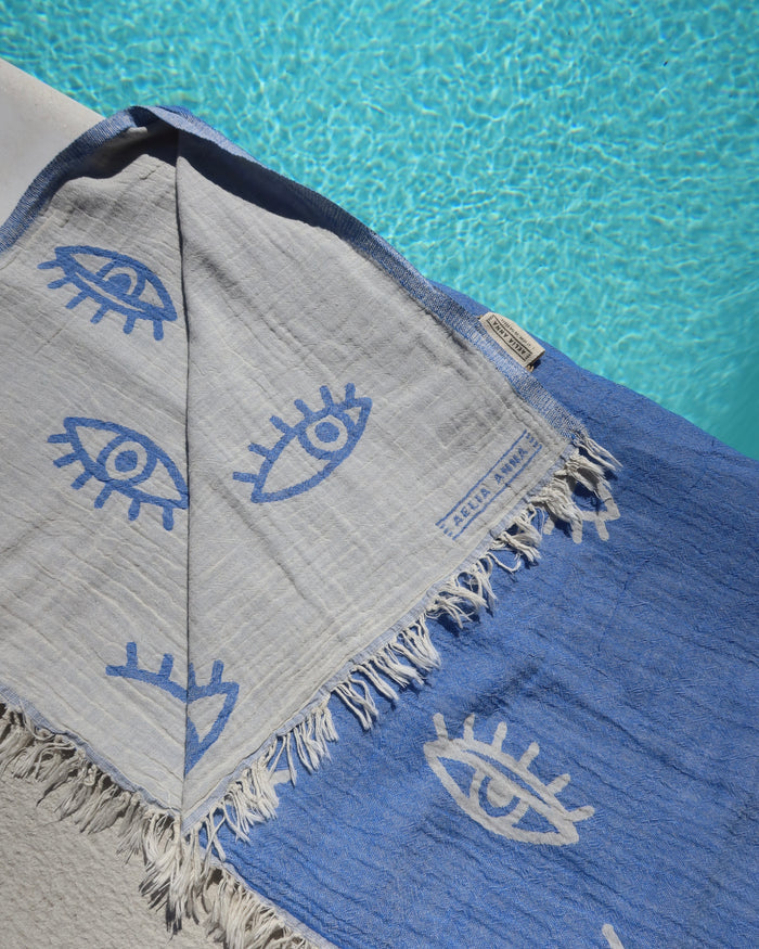 Blue cotton towel with white evil eye patterns  and a reversible white and blue evil eye print on a poolside