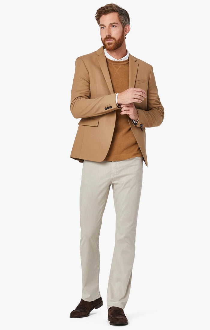 A man with light brown hair and a beard wears a tan blazer, brown sweater, white collared shirt, 34 HERITAGE - CHARISMA RELAXED STRAIGHT LEG PANT IN DAWN TWILL by 34 HERITAGE, and brown shoes against a plain white background.