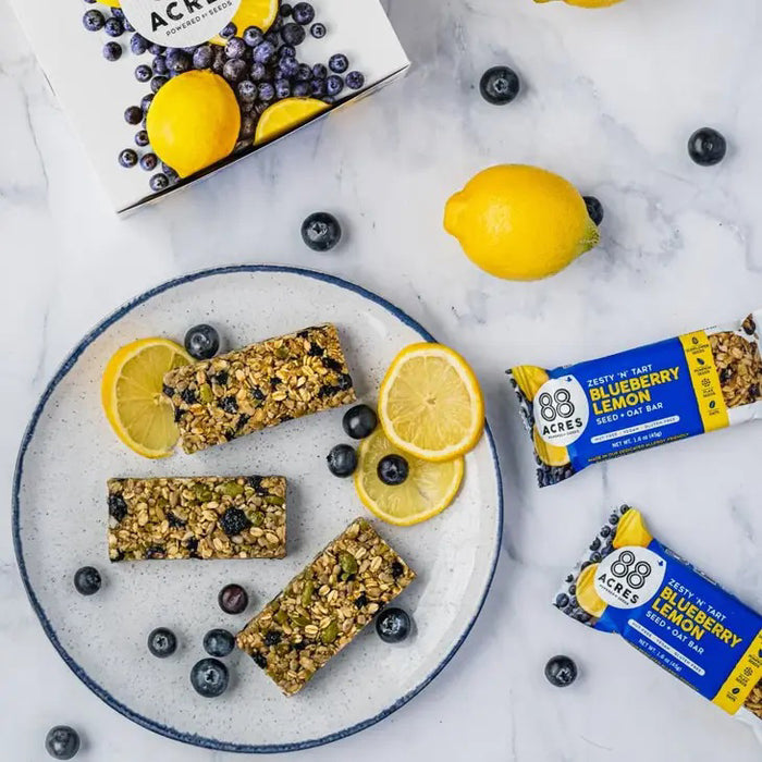 An 88 ACRES - BLUEBERRY LEMON SEED OAT BAR rests in its packaging on a white wooden surface, displaying images of blueberries and lemons. The gluten-free, vegan bar from 88 ACRES is nut-free, nutritious, and delicious.