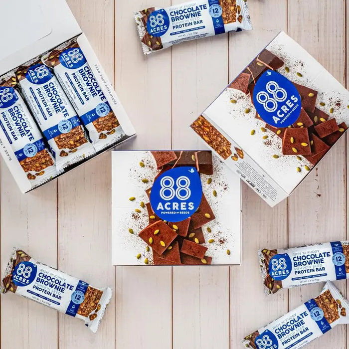 An 88 ACRES Fudge Brownie High Protein Bar is displayed on a white background. The wrapper, predominantly white with blue accents, showcases images of the bar and its ingredients, emphasizing that it is a nut-free protein bar from 88 ACRES.