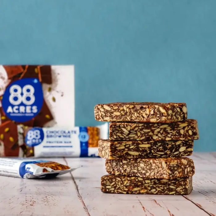 An 88 ACRES Fudge Brownie High Protein Bar is displayed on a white background. The wrapper, predominantly white with blue accents, showcases images of the bar and its ingredients, emphasizing that it is a nut-free protein bar from 88 ACRES.