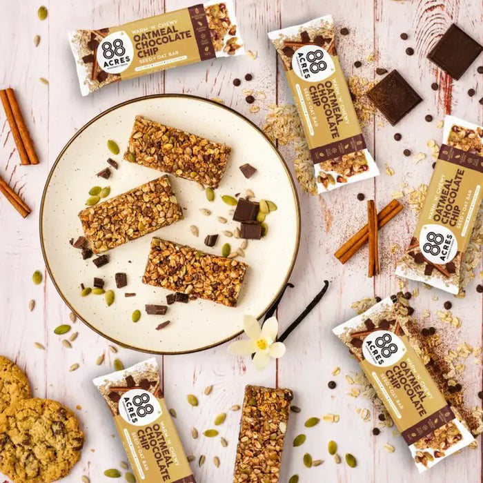 An 88 ACRES - OATMEAL CHOCOLATE CHIP SEED OAT BAR is shown on a light wooden surface, its brown and white wrapper featuring oats, chocolate chips, and Australian sea salt for a chewy cookie bar experience.