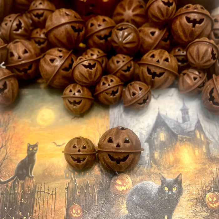 A set of rusty brown metal pumpkin bells in two varied sizes with cute carved jack-o'-lantern faces, arranged on a Halloween-themed printed background featuring a haunted house, pumpkins, and black cats.