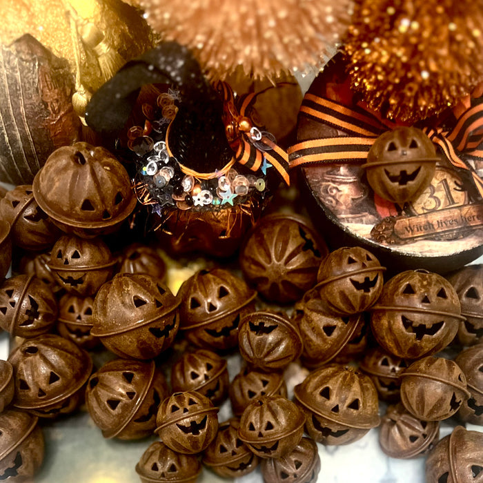 A collection of carved jack-o'-lantern metal bells in a warm rusty brown tone, arranged with Halloween-themed decorative props including a decorated pumpkin, orange-and-black striped ribbon, and a paper mache Halloween box in the background.