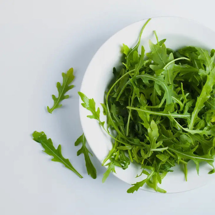 BELLA CUCINA - ARUGULA AND PINE NUT PESTO by BELLA CUCINA is showcased in a white bowl on a light surface, surrounded by fresh arugula leaves and toasted pine nuts for garnish.