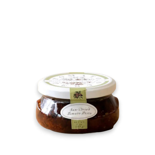 A jar of BELLA CUCINA - SUN-DRIED TOMATO PESTO is displayed against a plain backdrop. The jar has a white lid, labeled with the product name and weight (6 oz / 183 g). The rich, dark red pesto inside is thick and visible through the transparent glass jar, highlighting the quality of vine-ripened tomatoes used.