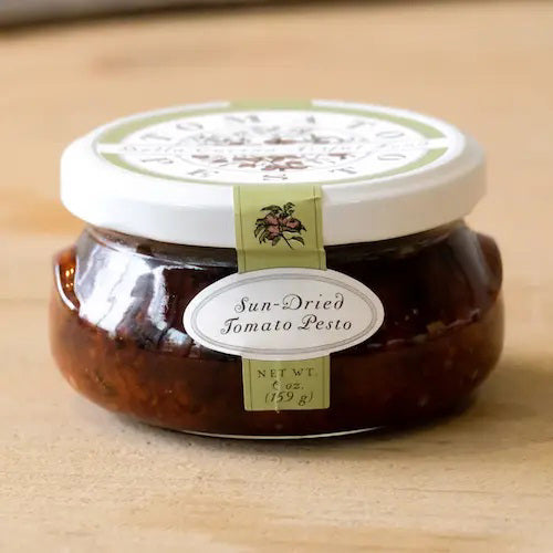 A jar of BELLA CUCINA - SUN-DRIED TOMATO PESTO is displayed against a plain backdrop. The jar has a white lid, labeled with the product name and weight (6 oz / 183 g). The rich, dark red pesto inside is thick and visible through the transparent glass jar, highlighting the quality of vine-ripened tomatoes used.