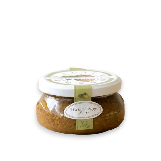 BELLA CUCINA's WALNUT SAGE PESTO is presented in a glass jar with a white lid, adorned with a decorative label showcasing a small bee illustration. Filled with finely blended pesto, it's rich in extra virgin olive oil and earthy walnuts, set against a plain white background.