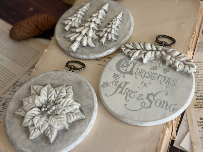 Three round gray decorative wall plaques with Christmas designs, displayed on vintage book pages. One plaque features a poinsettia, another has Christmas trees, and the third reads 'Christmas in Art & Song' with a wreath at the top.