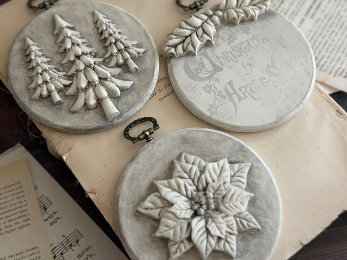 Three round gray decorative ornaments with raised holiday designs, including Christmas trees, holly, and a poinsettia, arranged on vintage printed paper.