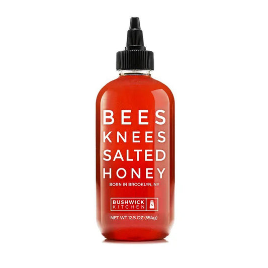 A 12.5 oz bottle of BUSHWICK KITCHEN - 'BEES KNEES' SALTED HONEY features a black twist cap and bright red label, blending wildflower honey with sea salt for a deliciously balanced sweet and savory flavor.