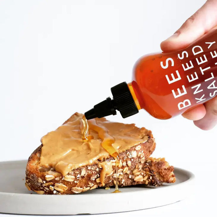 A 12.5 oz bottle of BUSHWICK KITCHEN - 'BEES KNEES' SALTED HONEY features a black twist cap and bright red label, blending wildflower honey with sea salt for a deliciously balanced sweet and savory flavor.
