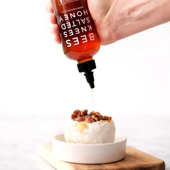 A 12.5 oz bottle of BUSHWICK KITCHEN - 'BEES KNEES' SALTED HONEY features a black twist cap and bright red label, blending wildflower honey with sea salt for a deliciously balanced sweet and savory flavor.