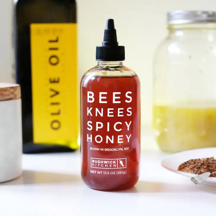 A 12.5 oz (354g) bottle of BUSHWICK KITCHEN - 'BEES KNEES' SPICY HONEY by BUSHWICK KITCHEN features an amber-colored honey visible through the clear container and a black twist cap. The label proudly states, "Born in Brooklyn, NY." This product is ideal for any honey gift set or for those who enjoy wildflower honeys with a spicy kick.