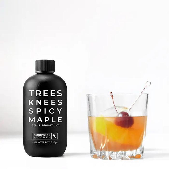 A 12.5 oz (354g) black bottle of BUSHWICK KITCHEN - 'TREES KNEES' SPICY MAPLE SYRUP features white text, a Brooklyn skyline graphic, and a black cap, containing habanero-infused spicy maple syrup.