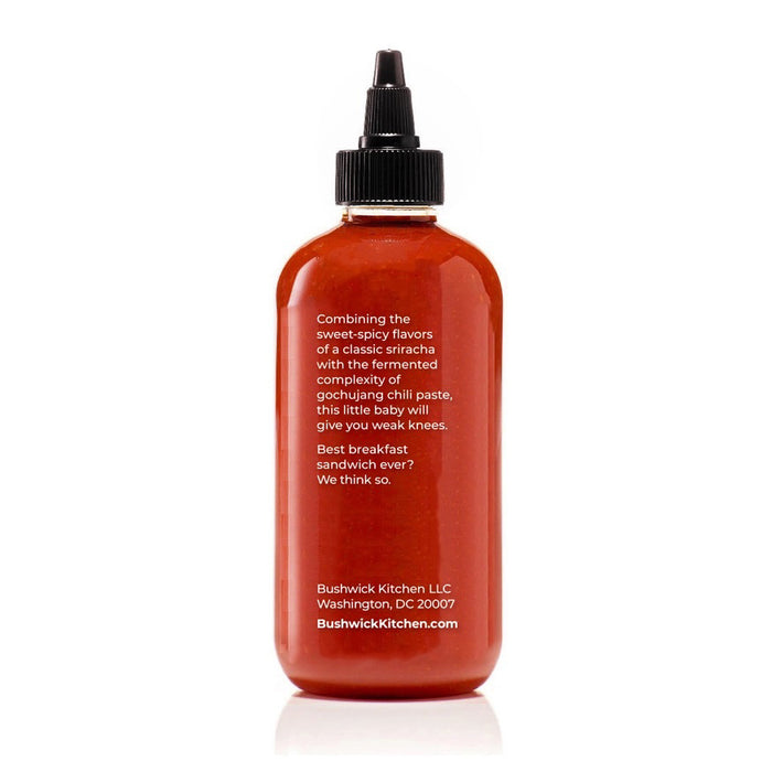 A clear bottle of BUSHWICK KITCHEN - 'WEAK KNEES' GOCHUJANG SRIRACHA hot sauce with a black squeeze cap, vibrant red chile paste, and label showing the name, "Born in Brooklyn, NY." Net wt. 10.5 oz (298g).