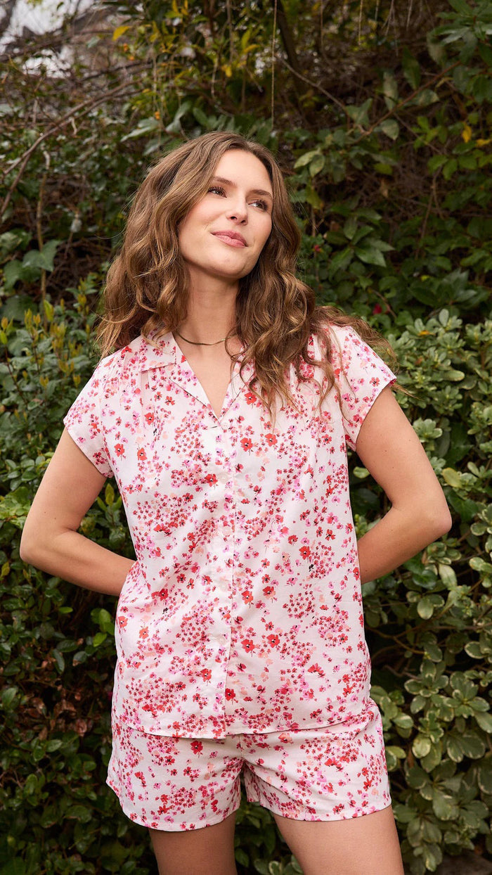 A smiling woman stands barefoot against a beige background, wearing the CAT'S PAJAMAS - PETITE ROSETTE VOILE SHORT SET by CAT'S PAJAMAS—a pink and white floral cotton voile pajama top and matching shorts—ideal for lightweight women's sleepwear.